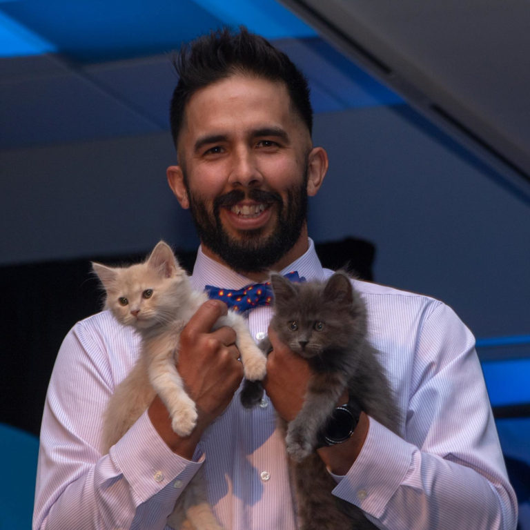 A smiling man with black hair and a beard holds two small kittens in his hands