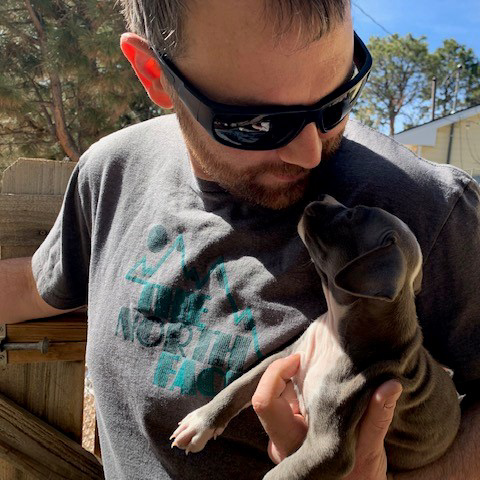 A man with a beard wearing sunglasses looks down at a small, gray puppy