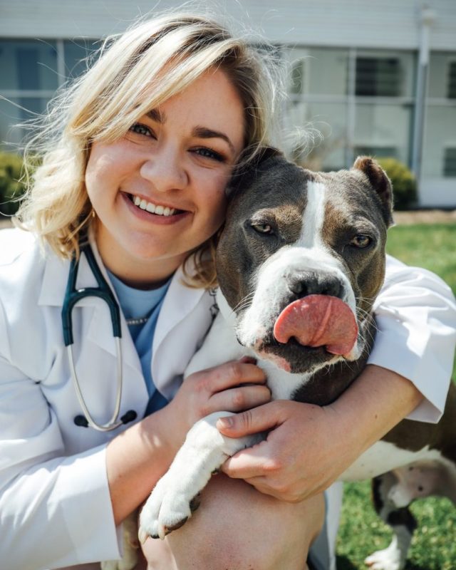 Blonde woman in white lab coat hugging a gray and white pit bull