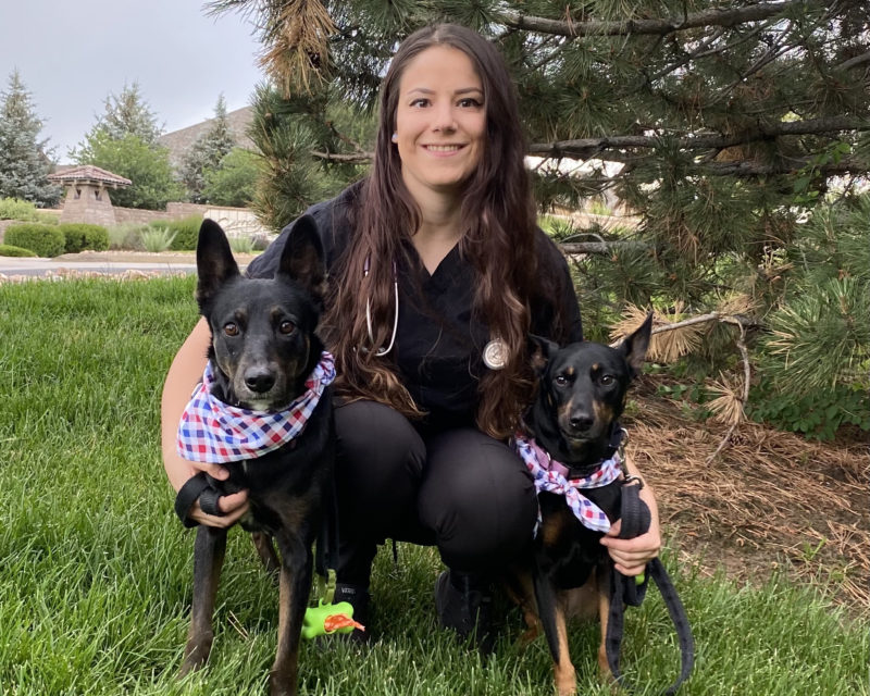 A white woman with dark brown hair in her vet uniform with her two small black dogs