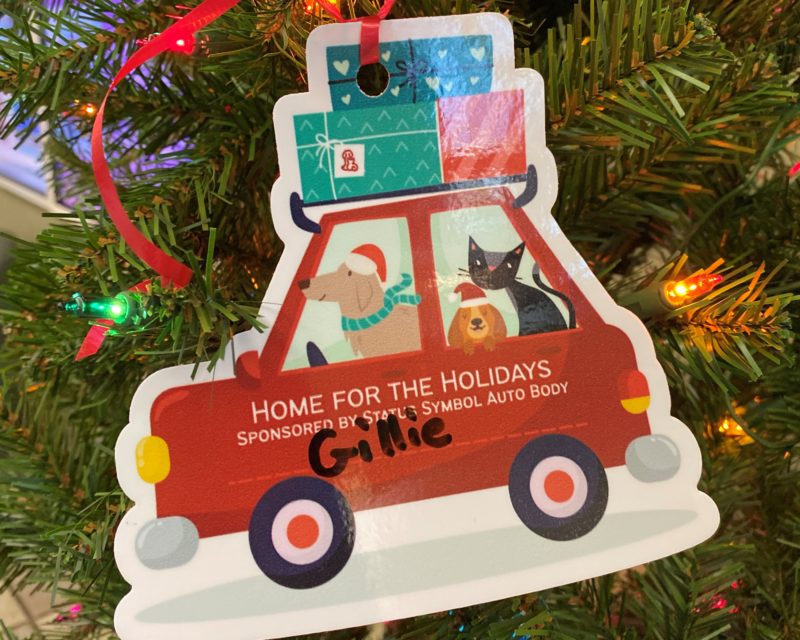 a car ornament with two dogs and a cat in the windows is hanging on a lit up Christmas tree