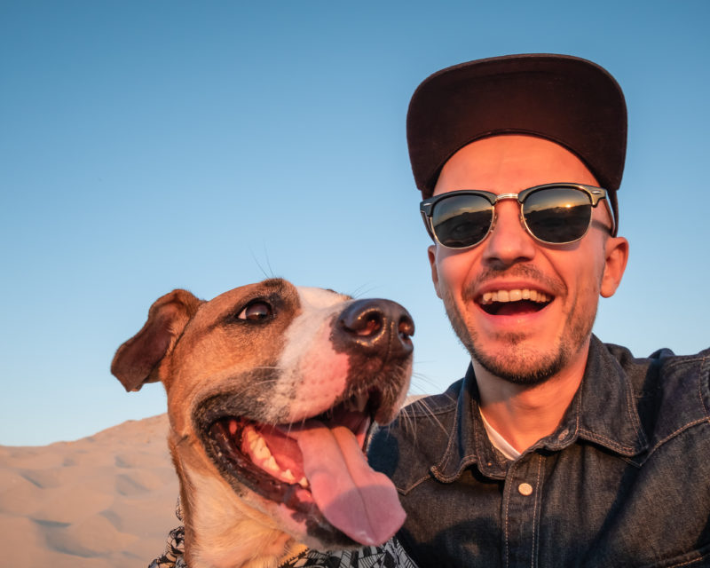 a white man in a baseball cap and sunglasses smiles next to his brown and white dog